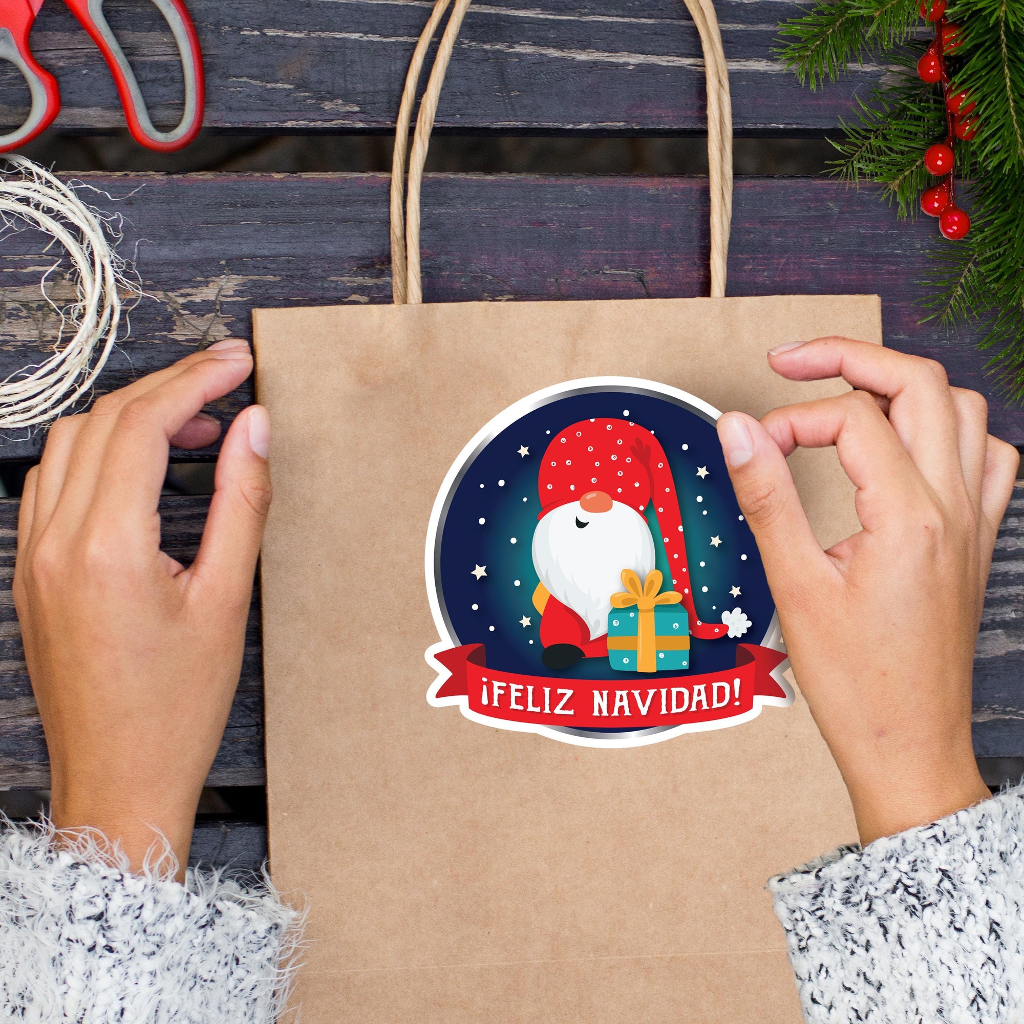 A person&#39;s hands holding a brown paper bag with a Christmas-themed sticker on it, featuring a gnome and the text &quot;Feliz Navid!&quot;