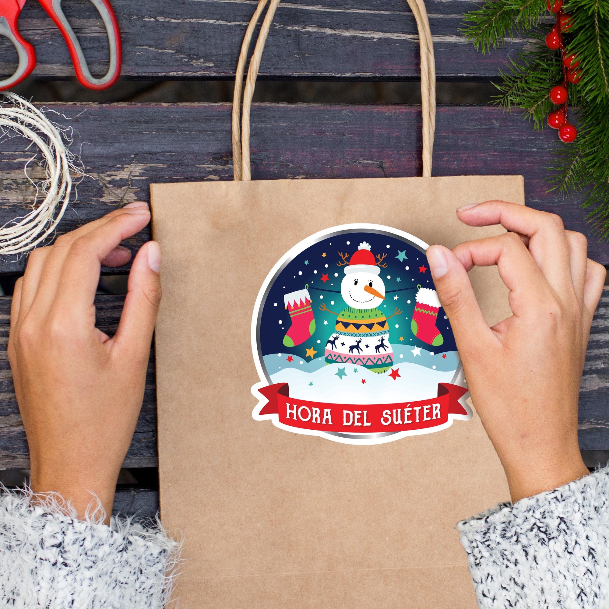 A person&#39;s hands are holding a brown paper bag with a festive snowman sticker on it, which features a red scarf, a snowman, and a red ribbon.