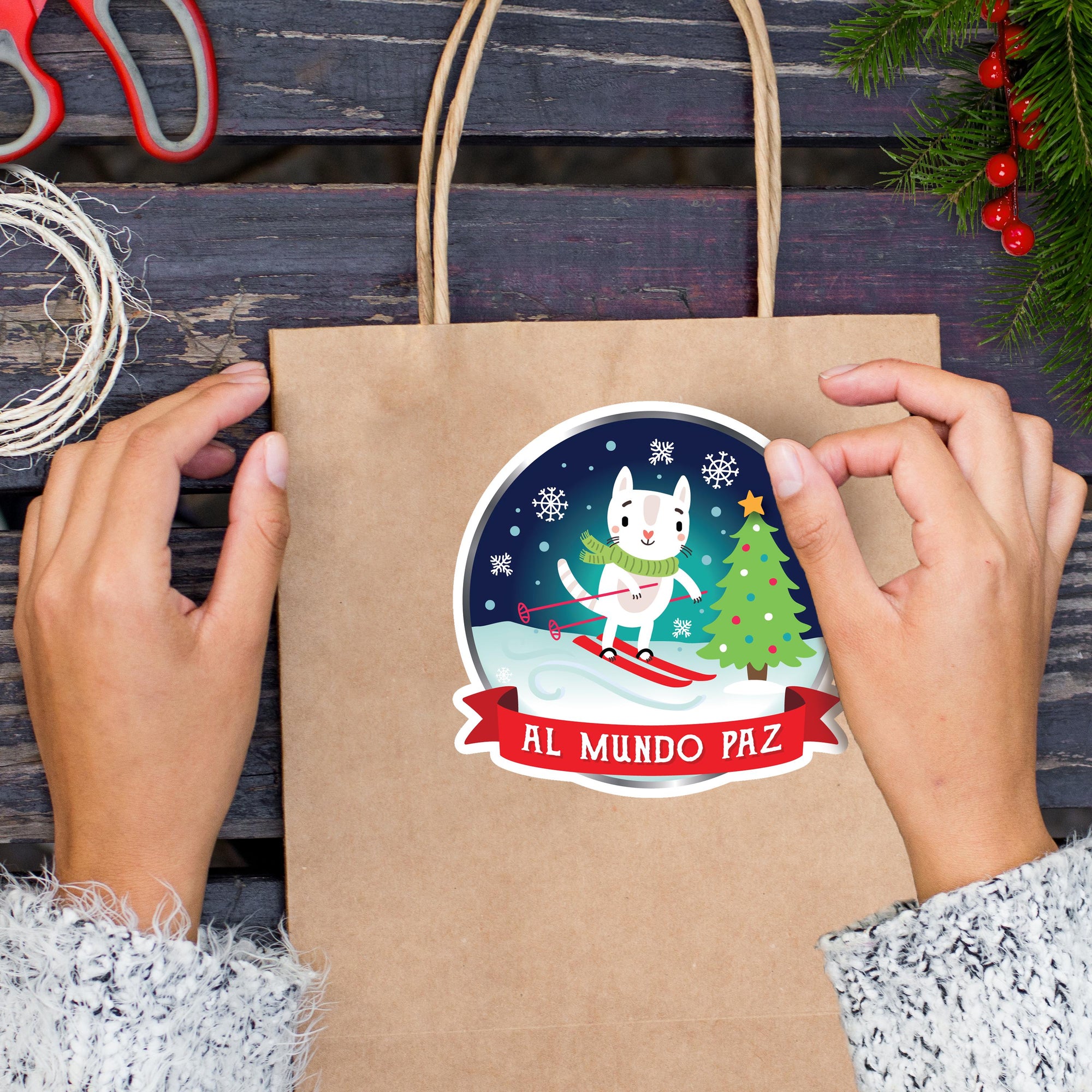A person&#39;s hands holding a brown paper bag with a sticker featuring a cartoon cat skiing on a snowy slope, with a Christmas tree and snowflakes in the background.