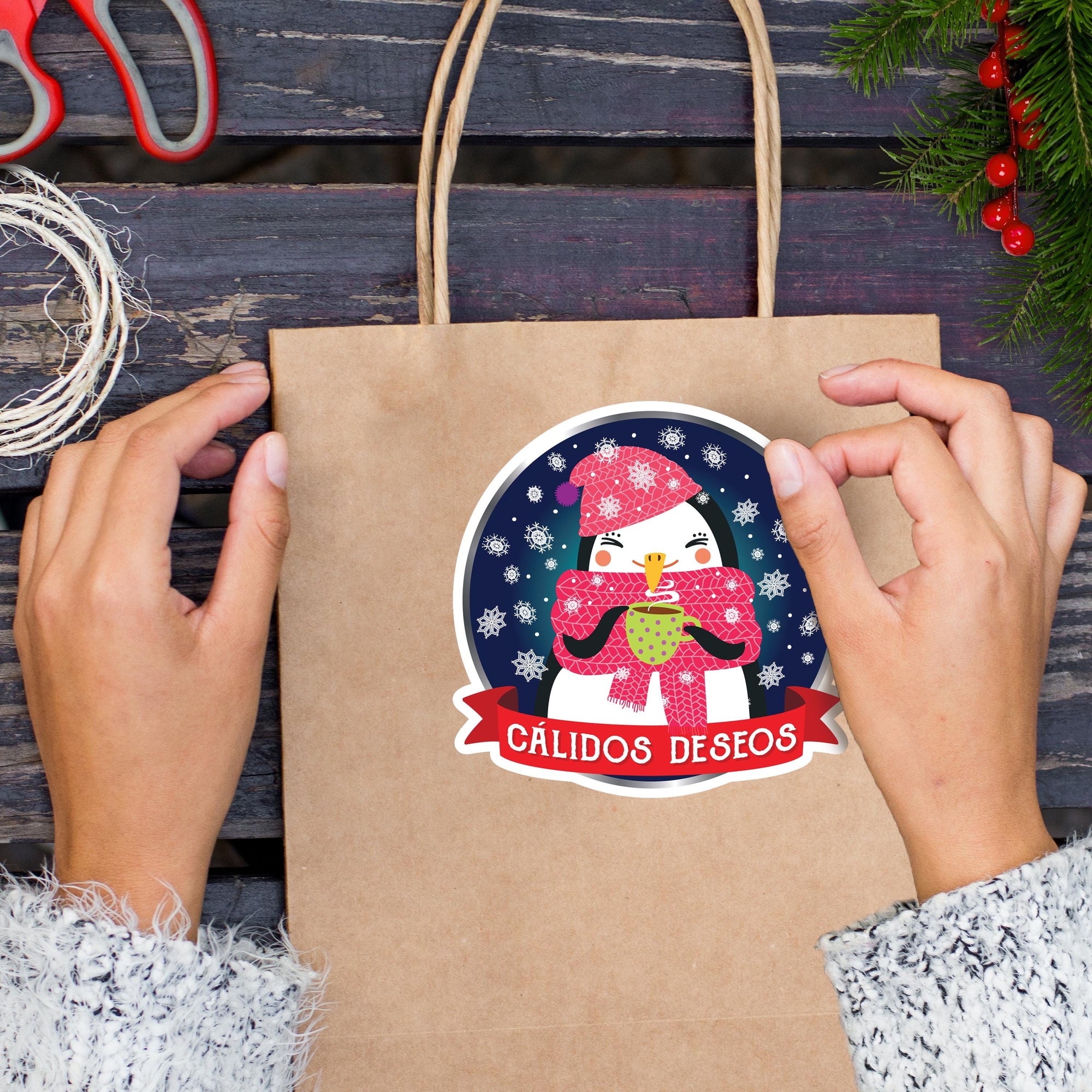 A person&#39;s hands holding a brown paper bag with a sticker featuring a cartoon penguin and the text &quot;CALIDOS DESEOS&quot; (Spanish for &quot;Happy Christmas&quot;).