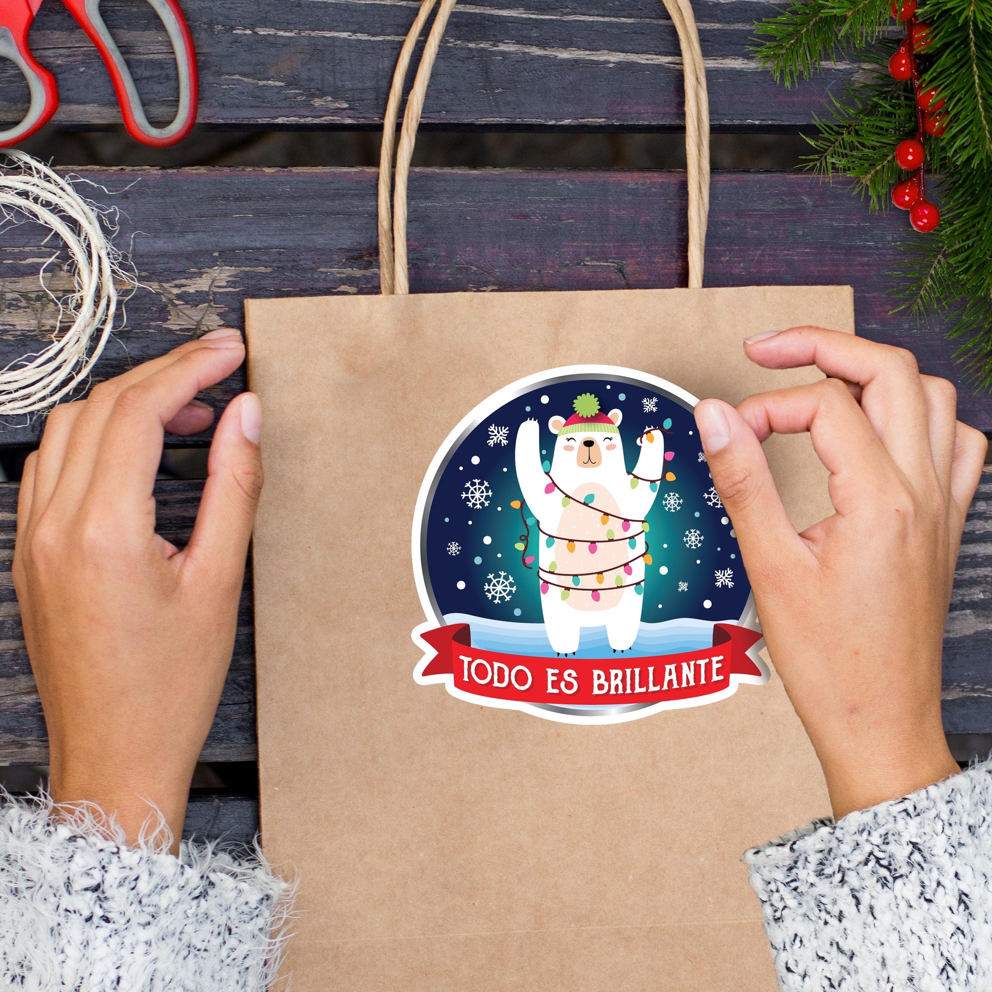 A person&#39;s hands are holding a brown paper bag with a sticker featuring a cartoon polar bear wearing a hat and surrounded by snowflakes, with the text &quot;TODO ES BRILLANTE&quot; (which means &quot;Do it yourself&quot; in Spanish).