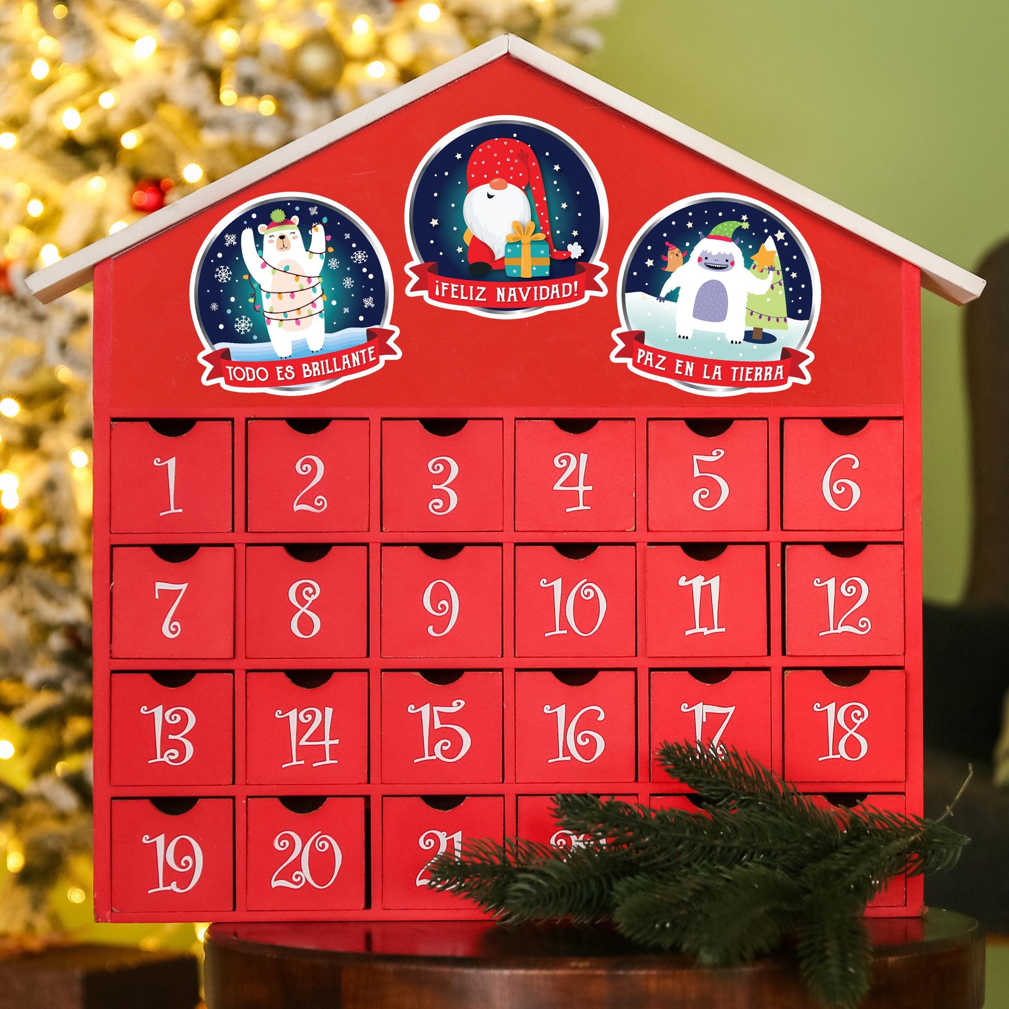 A red advent calendar with numbered drawers, featuring illustrations of Santa Claus, a reindeer, and a snowman. The calendar is placed on a table with a pine branch and a Christmas tree in the background.