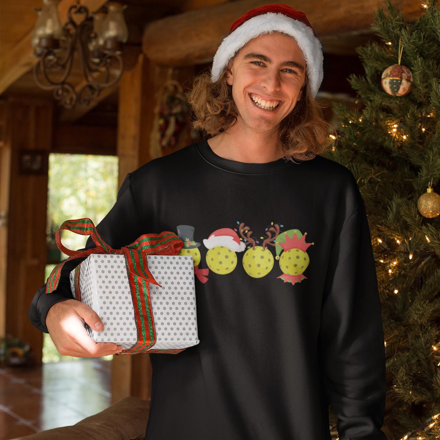 A smiling man wearing a black sweater with a graphic of three yellow tennis balls and a red hat holding a gift box.