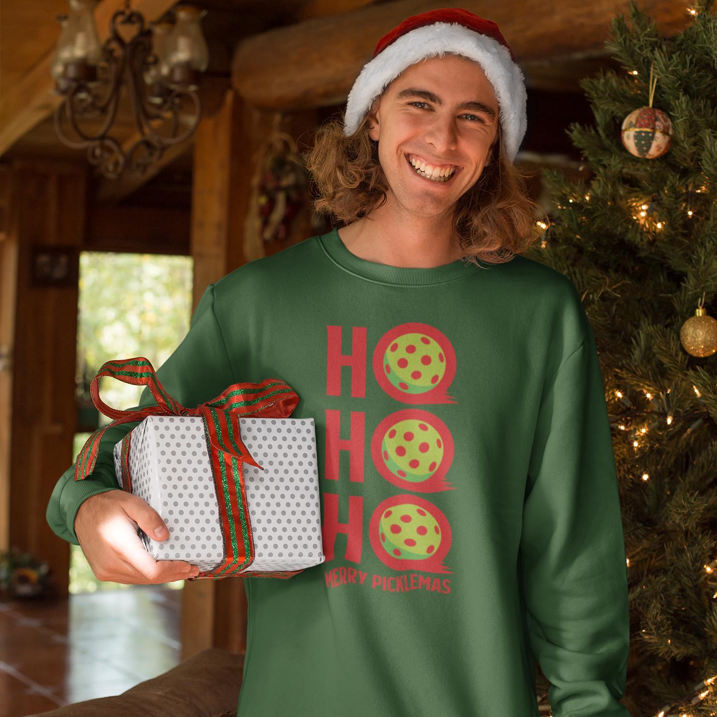 A smiling man wearing a Santa hat and a green sweater with red and yellow polka dots holding a gift box in front of a Christmas tree.