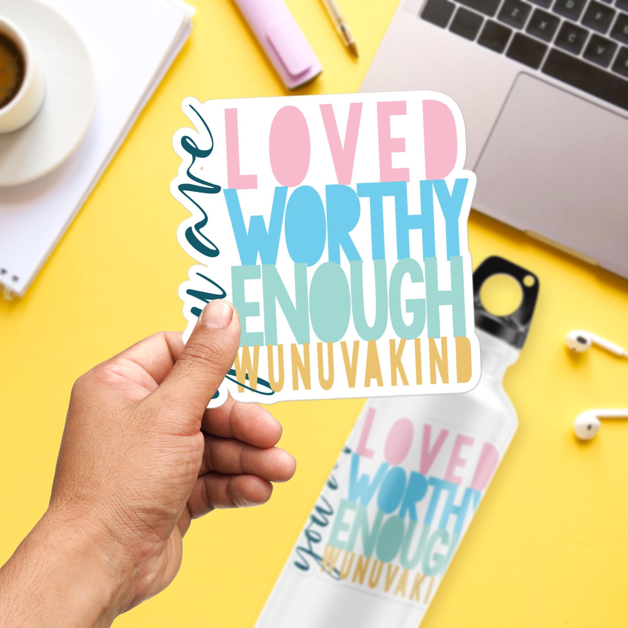 A person's hand holding a colorful sticker with the text "LOVED WORTHY ENOUGH WUNUVAKIND" against a yellow background.