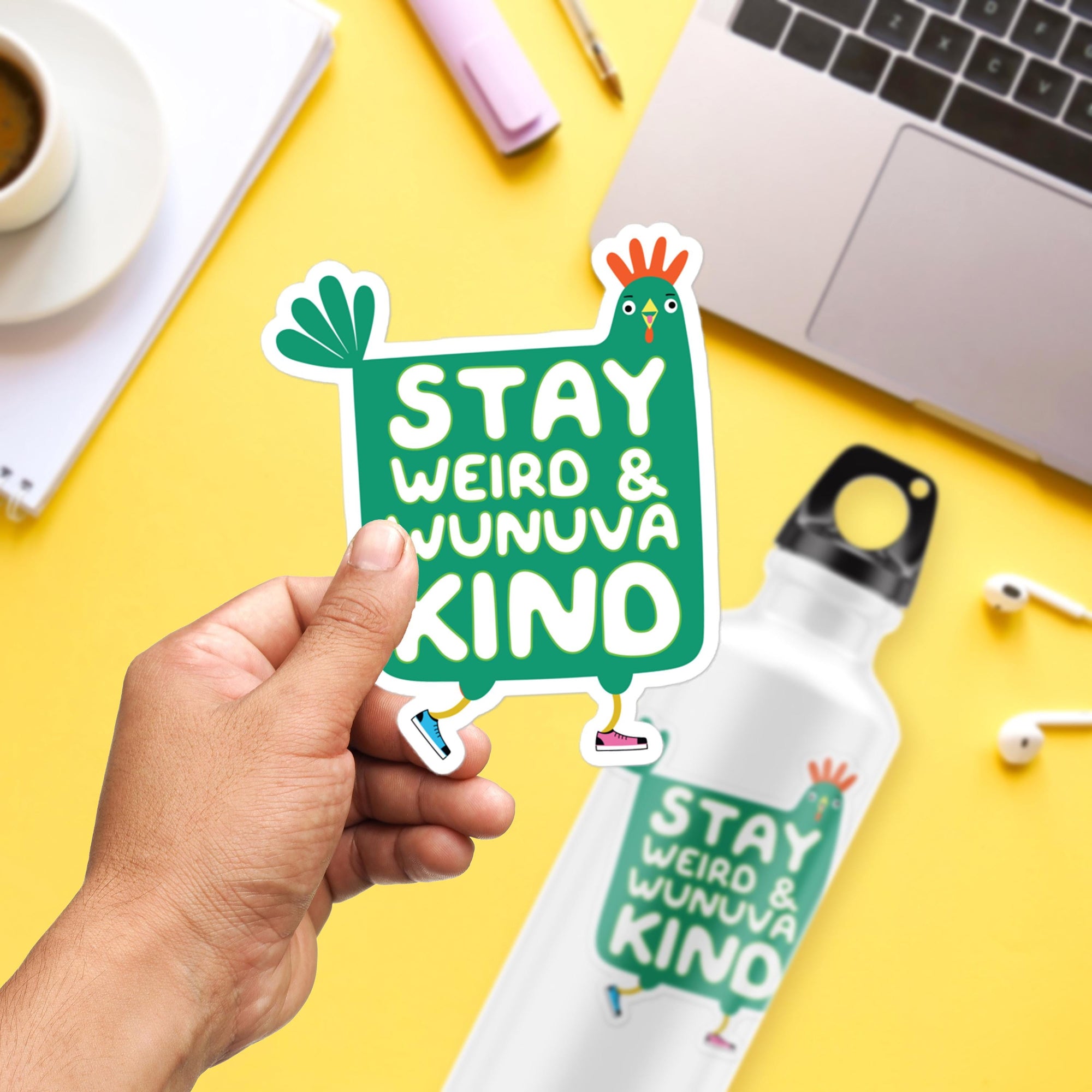 A person's hand holding a sticker with the text "STAY WEIRD & WUNUVA KIND" against a yellow background with various objects such as a laptop, headphones, and a water bottle.