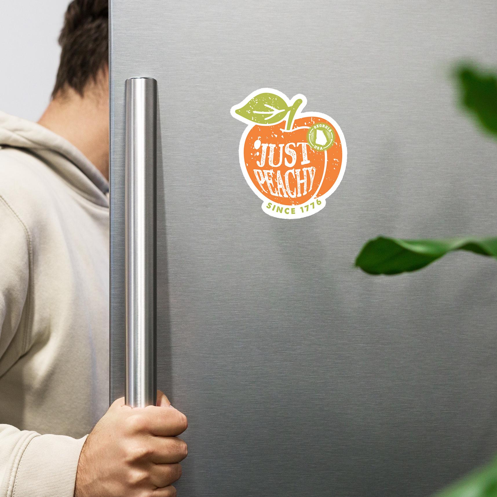 A person is holding a metal handle on a refrigerator door with a sticker of an orange peach and the text "Just Peachy" on it.