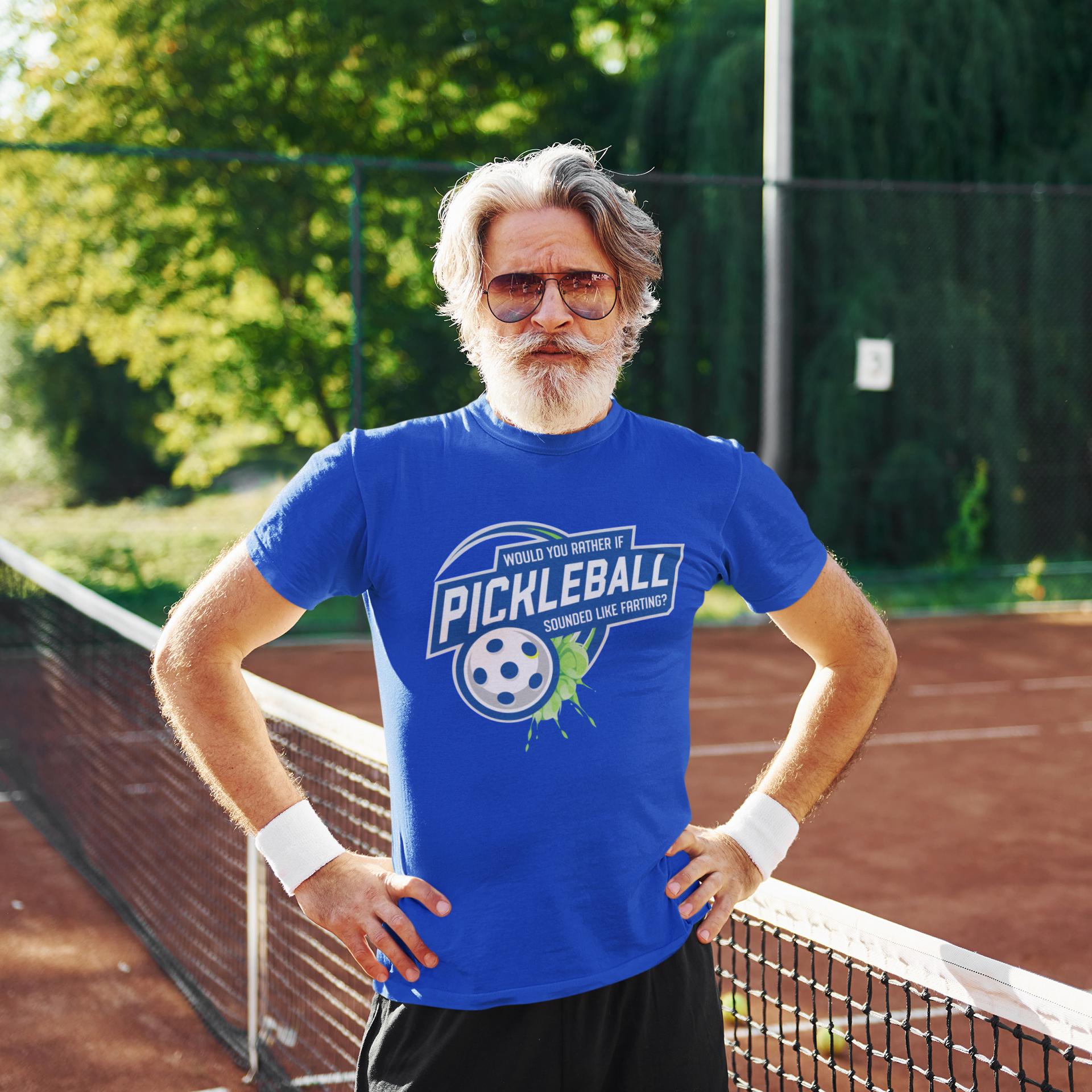 A man wearing a blue t-shirt with a humorous text and graphic stands on a tennis court, with trees and a fence visible in the background.