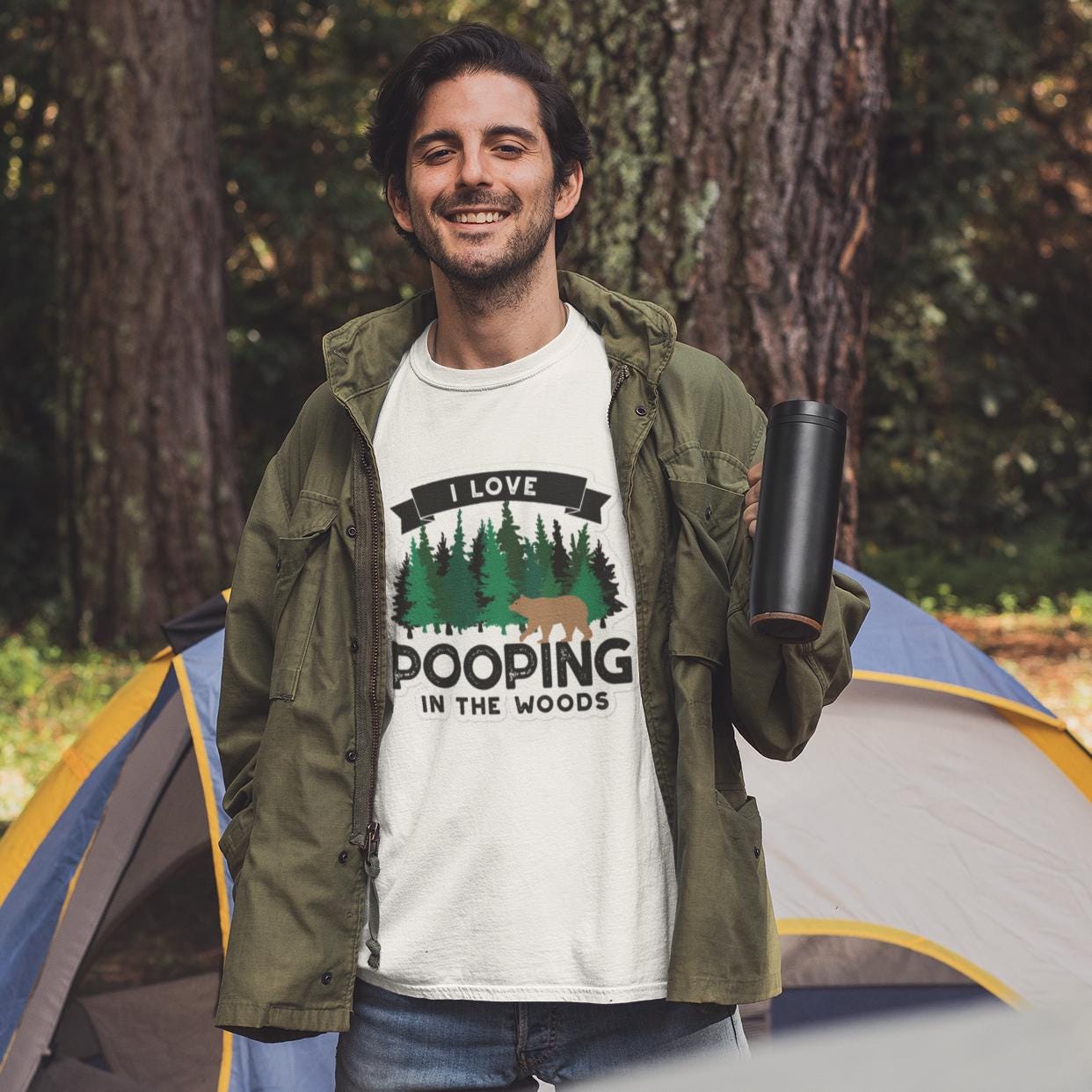 A man wearing a white t-shirt with a graphic design and holding a black object, standing in front of a tent in a forested area.