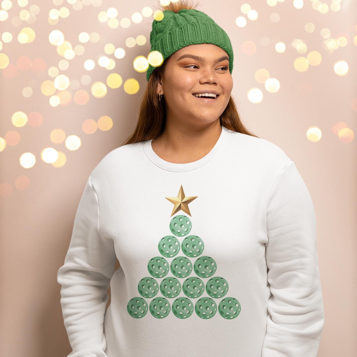 A woman wearing a white sweater with a Christmas tree design and a green beanie is smiling and looking to the side.