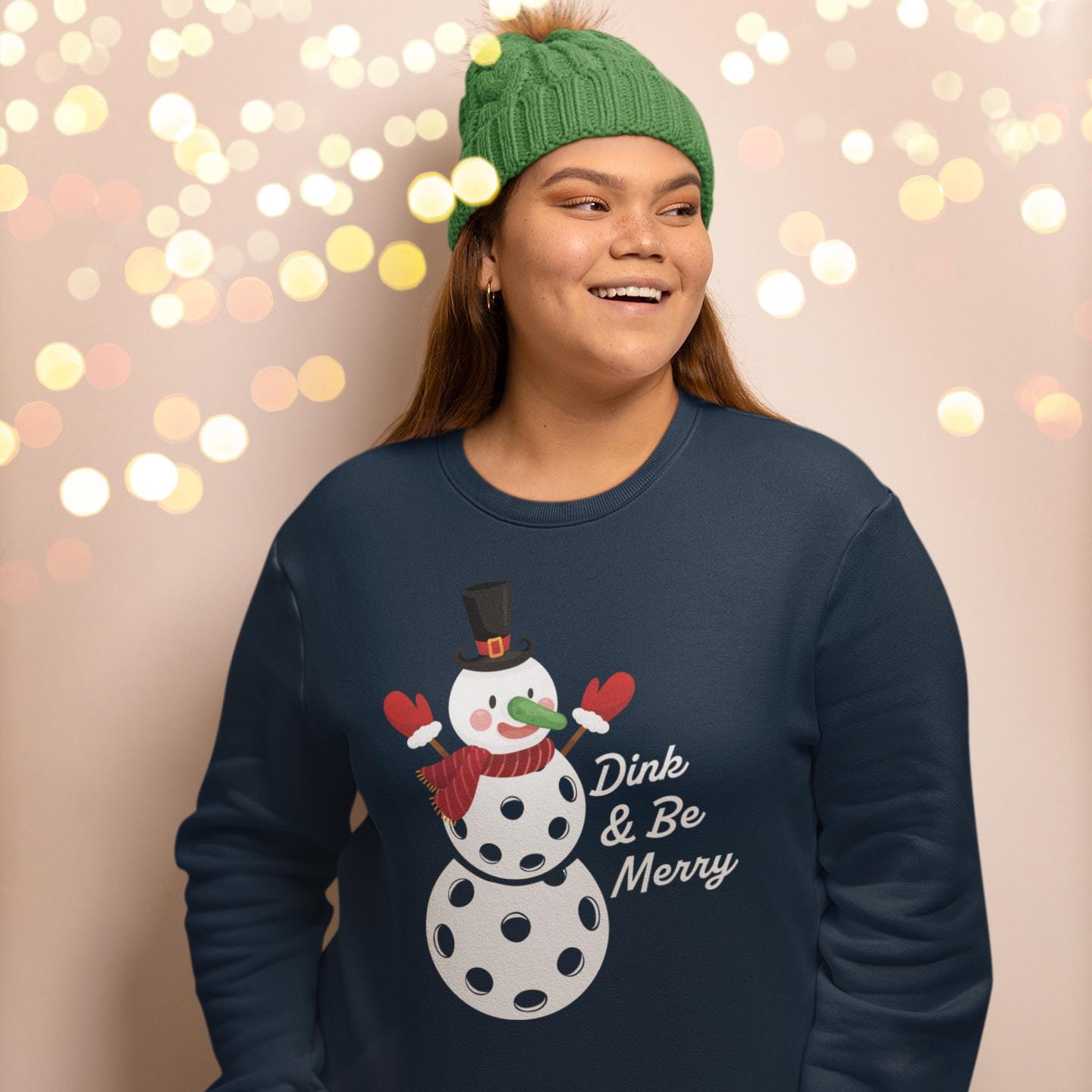 A woman wearing a green knit cap and a navy blue sweatshirt with a snowman graphic and the text "Dink & Be Merry" is smiling and looking to the side.