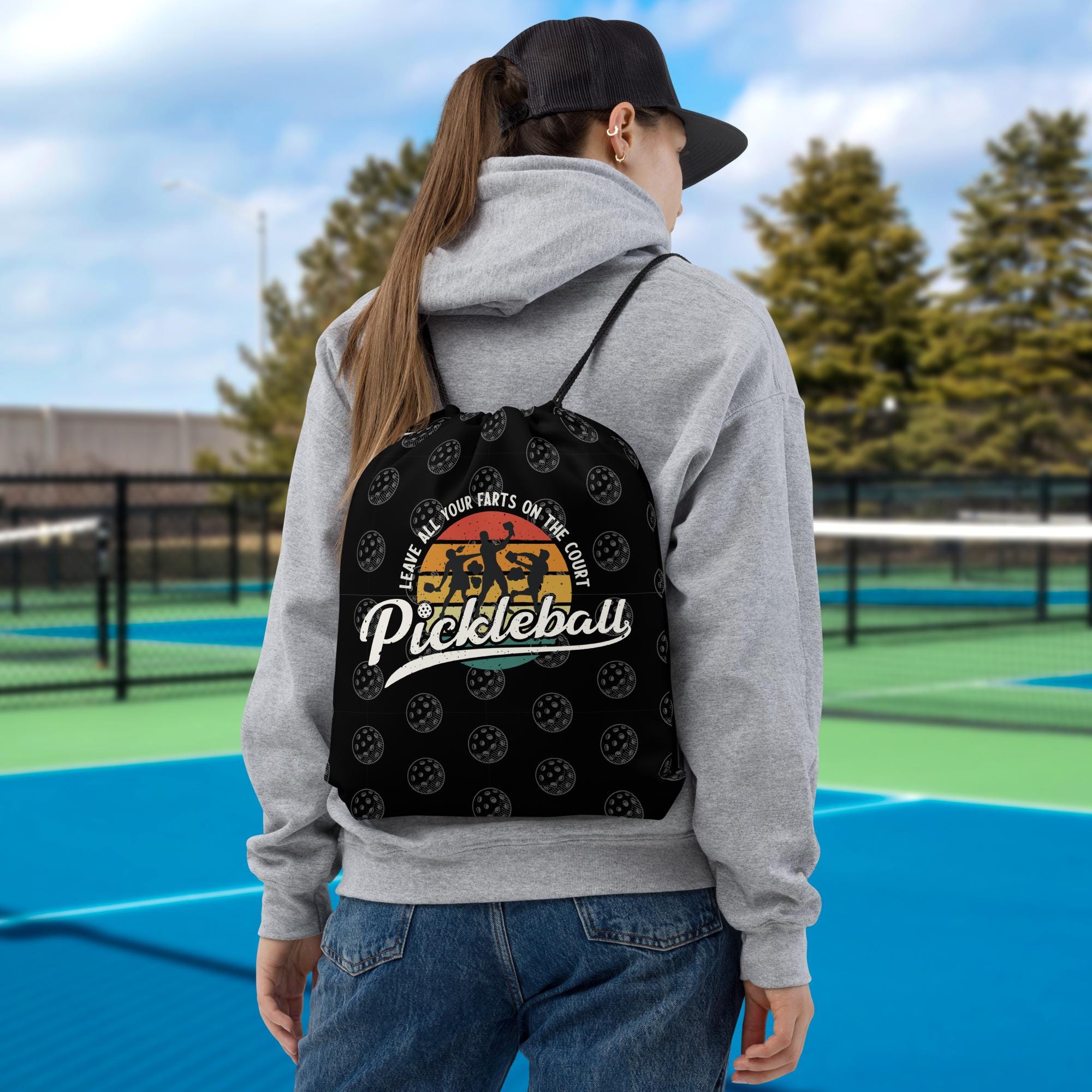 A person wearing a gray hoodie and a black baseball cap is standing on a tennis court, with a black backpack featuring a pattern of tennis balls on it.