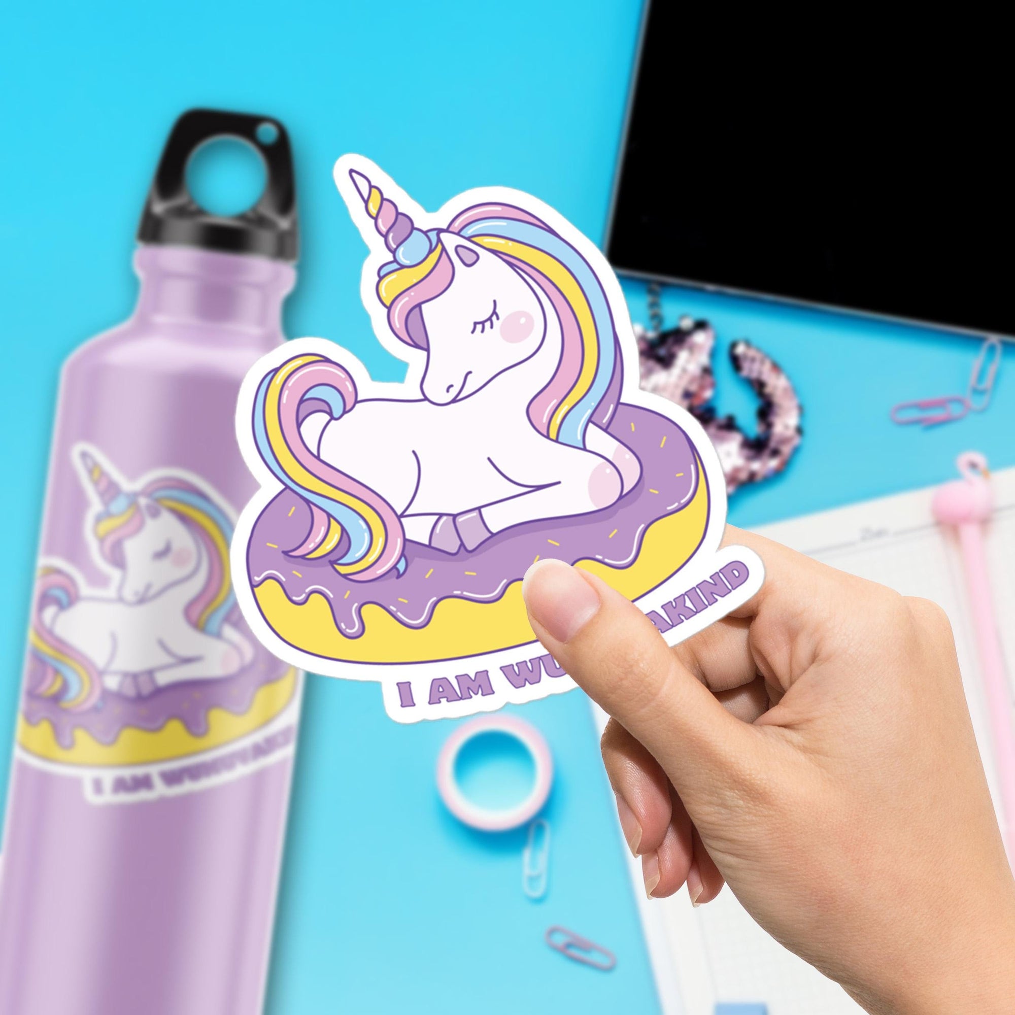 A hand is holding a sticker featuring a unicorn on a donut, with a water bottle and other office supplies visible in the background.