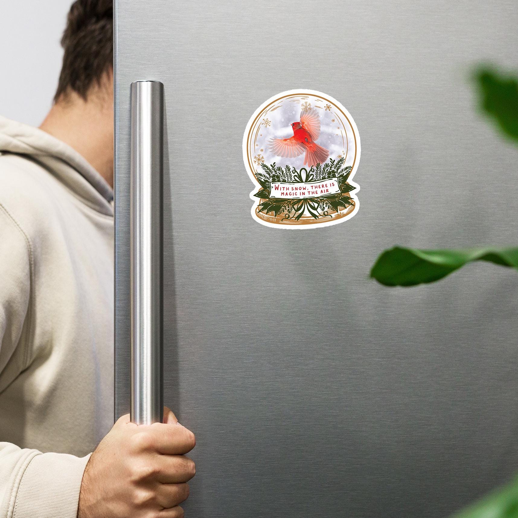 A person is holding a silver refrigerator handle, with a snow globe sticker featuring a red bird on the door.