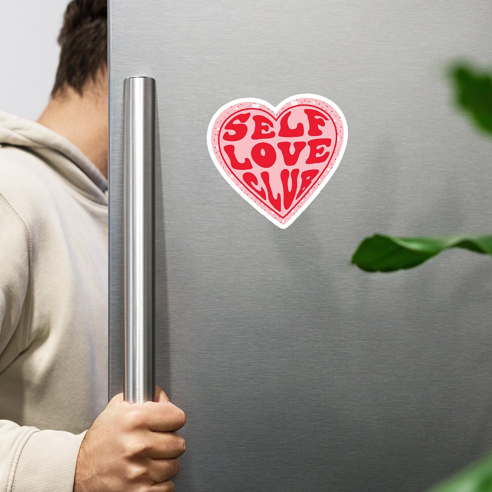 A person is holding a silver refrigerator handle, with a heart-shaped sticker on the refrigerator door that says "Self Love Club".