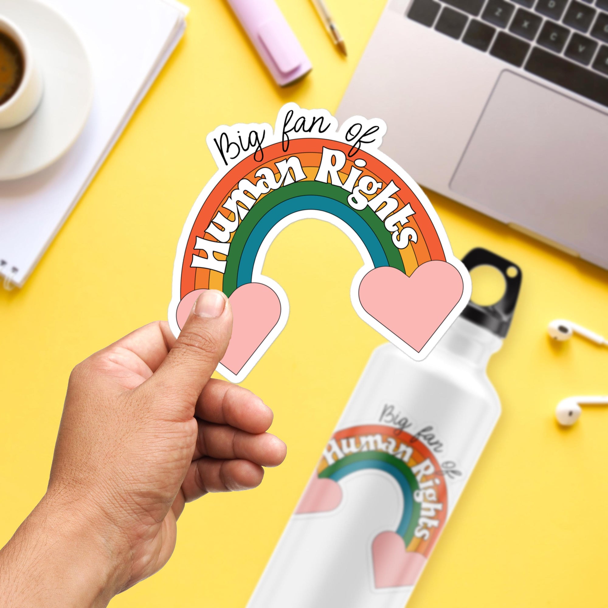 A person's hand holding a sticker with a rainbow and heart design, along with a laptop and other office supplies on a yellow background.
